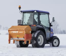 Produktbild: RAUCH Winterdienststreuer UKS, orange, montiert an Traktor mit Schneeketten im Wintereinsatz
