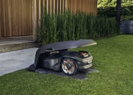 Lifestyle: Husqvarna® Mähroboter fährt aus schwarzer Garage auf Rasenfläche im Garten