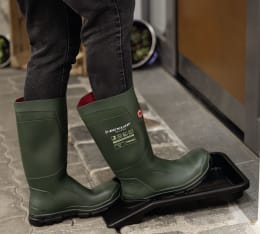 Lifestyle: Person nutzt Stiefelknecht mit Auffangwanne zum Ausziehen grüner Gummistiefel
