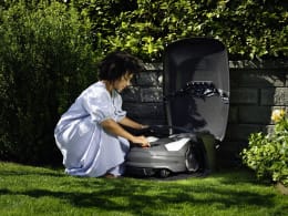 Lifestyle: Frau öffnet schwarze Mährobotergarage mit Mähroboter im Garten