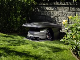 Lifestyle: Husqvarna® Mähroboter unter grauer Garage im Garten neben Steinmauer und Hecke