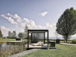 Lifestyle: Pergola mit Lamellenseitenwand im Garten, Sonnenliege auf Holzdeck, Teich im Hintergrund