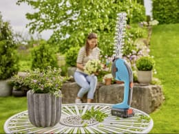 Lifestyle: GARDENA Akku-Grasschere PowerCut 20 auf Gartentisch, Frau im Hintergrund bei der Gartenarbeit