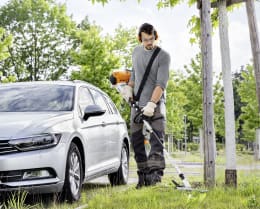 Lifestyle: Mann mit Gehörschutz trimmt Gras mit STIHL Benzin-Kombimotor KM 131 R neben Auto