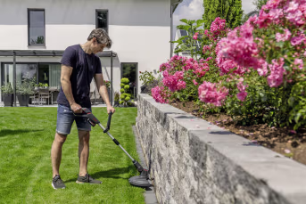 Lifestyle: Mann trimmt Rasenkante entlang einer Steinmauer mit Akku-Rasentrimmer im Garten