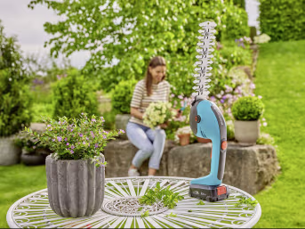 Lifestyle: GARDENA Akku-Grasschere PowerCut 20 auf Gartentisch, Frau im Hintergrund bei der Gartenarbeit