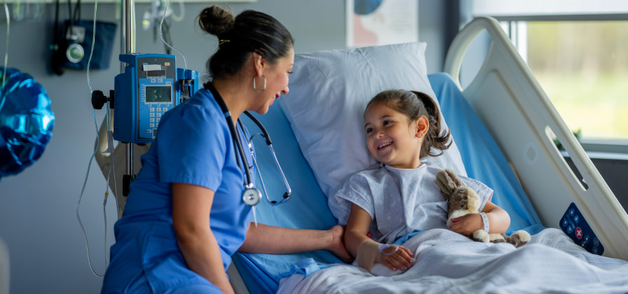 A cheerful nurse smiles with a child patient holding a stuffed animal in a hospital bed