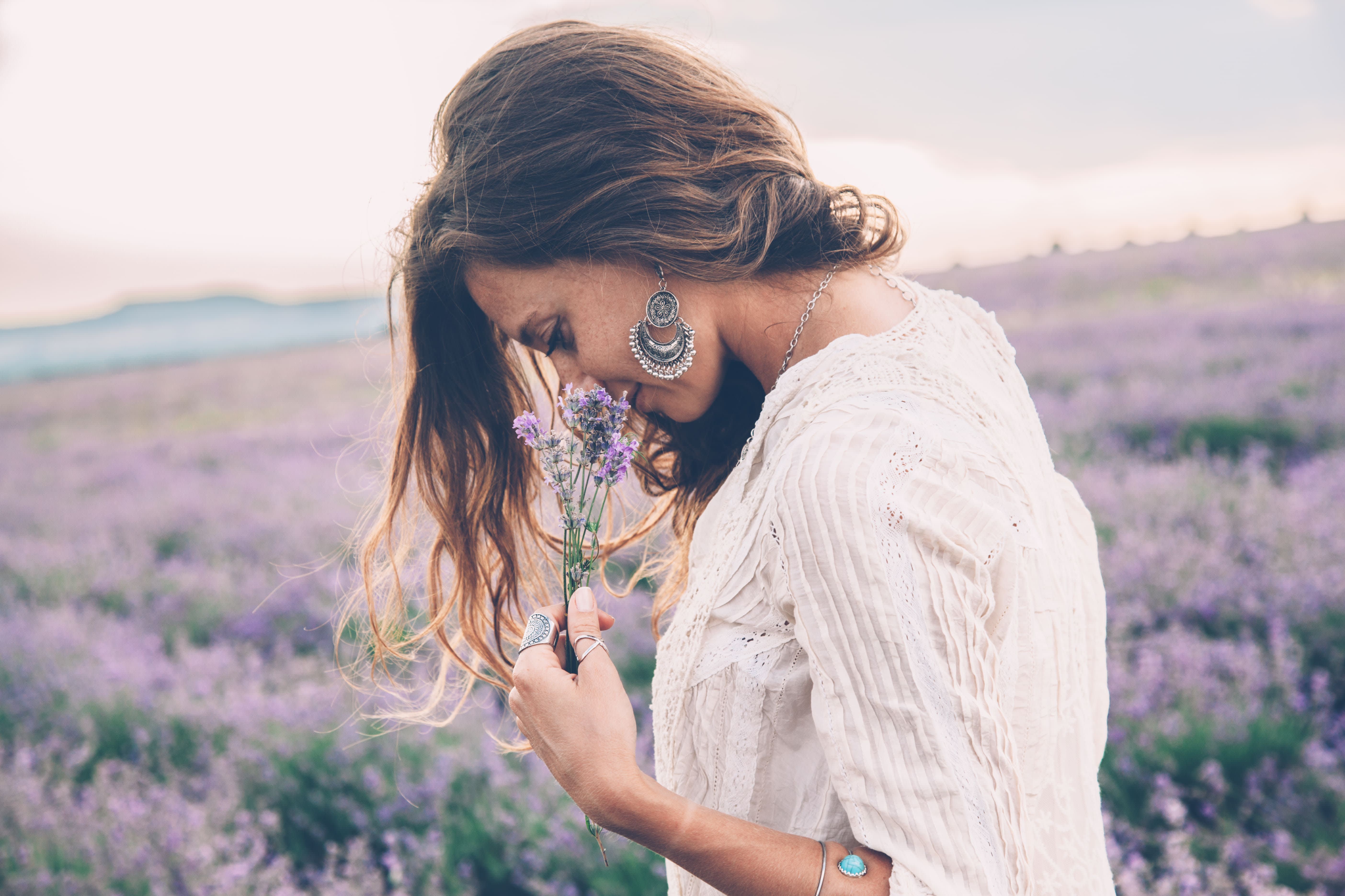 Mulher branca vestida de branco e com cabelos castanhos em um campo de Lavandas. Ela está cheirando flores de lavanda.