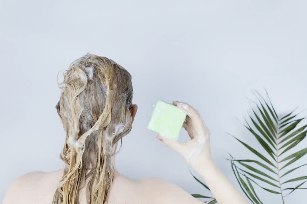 Imagem de pessoa com cabelos loiros ensaboados segurando um shampoo em barra