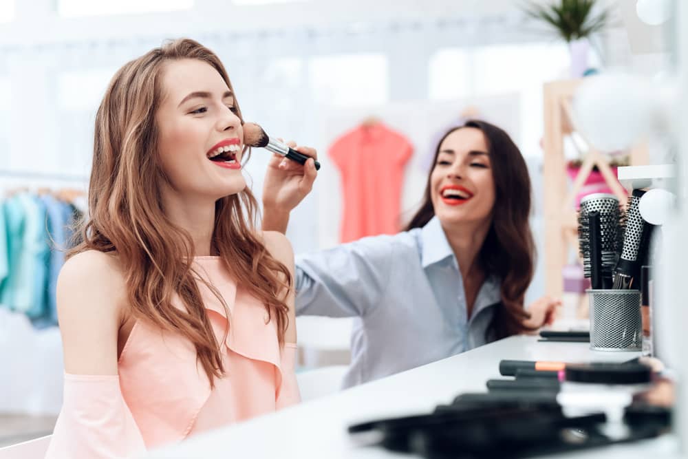 Imagens de duas mulheres sorrindo, elas estão se maquiando juntas e uma aplicar um produto no rosto da outra com um pincel de maquiagem