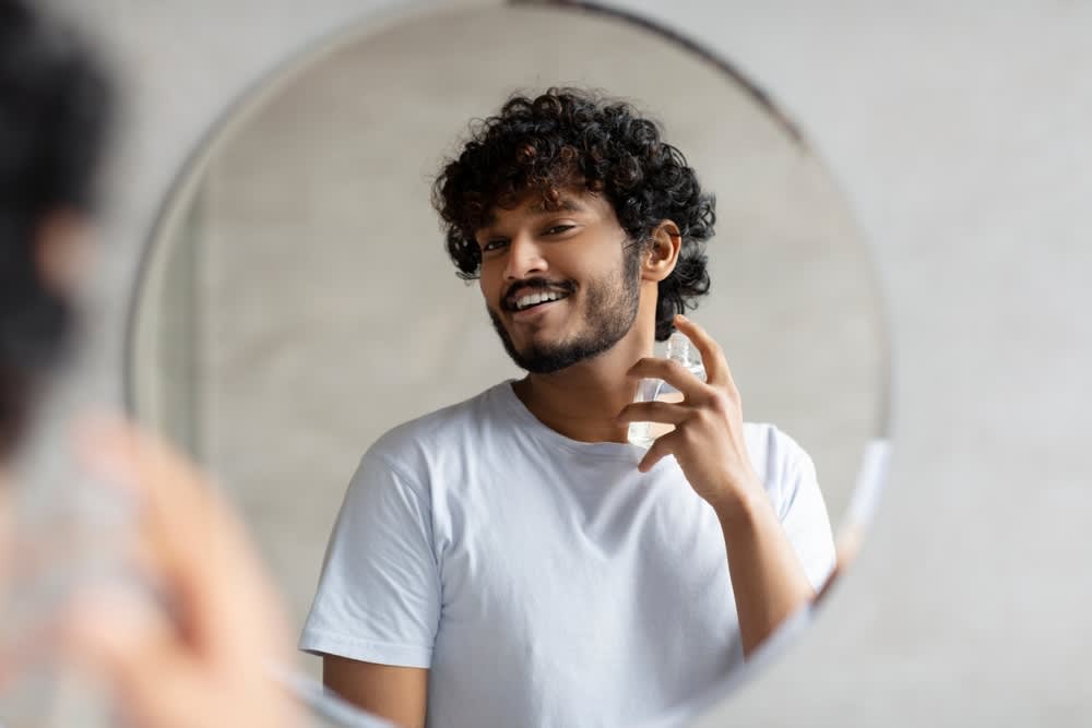 A imagem mostra um homem aplicando fragrância na pele em frente ao espelho