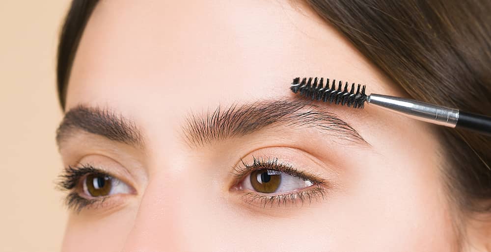 A imagem foca nos olhos de uma mulher branca. Ela está penteado a sobrancelha com um pincel escova