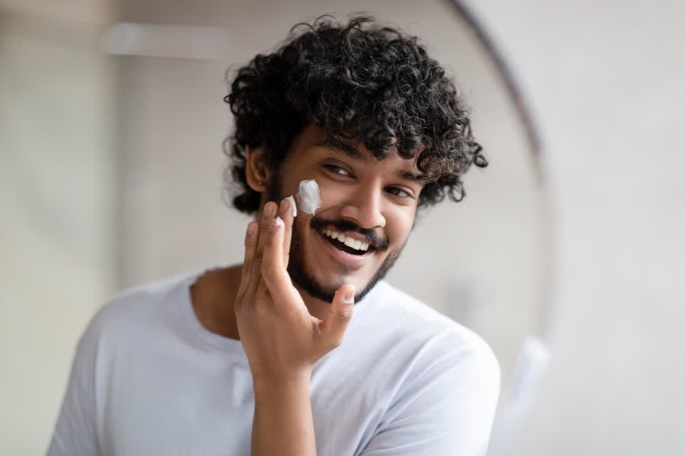 Homem negro sorrindo enquanto aplica creme facial no rosto para uma rotina de autocuidado