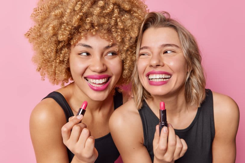 Duas mulheres, uma com longos cabelos crespos e loiros e outra com cabelo curto, liso e loiros. Ambas estão sorrindo enquanto aplicam batom