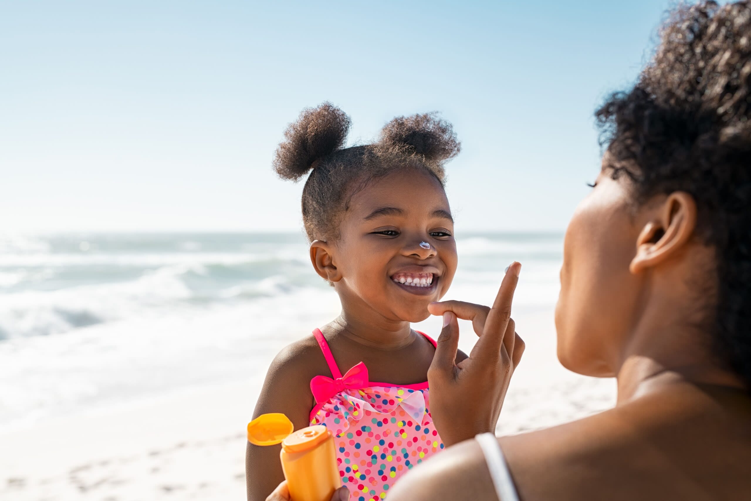 Imagem de mulher adulta e criança na praia. A mulher está passando protetor solar no rosto da criança