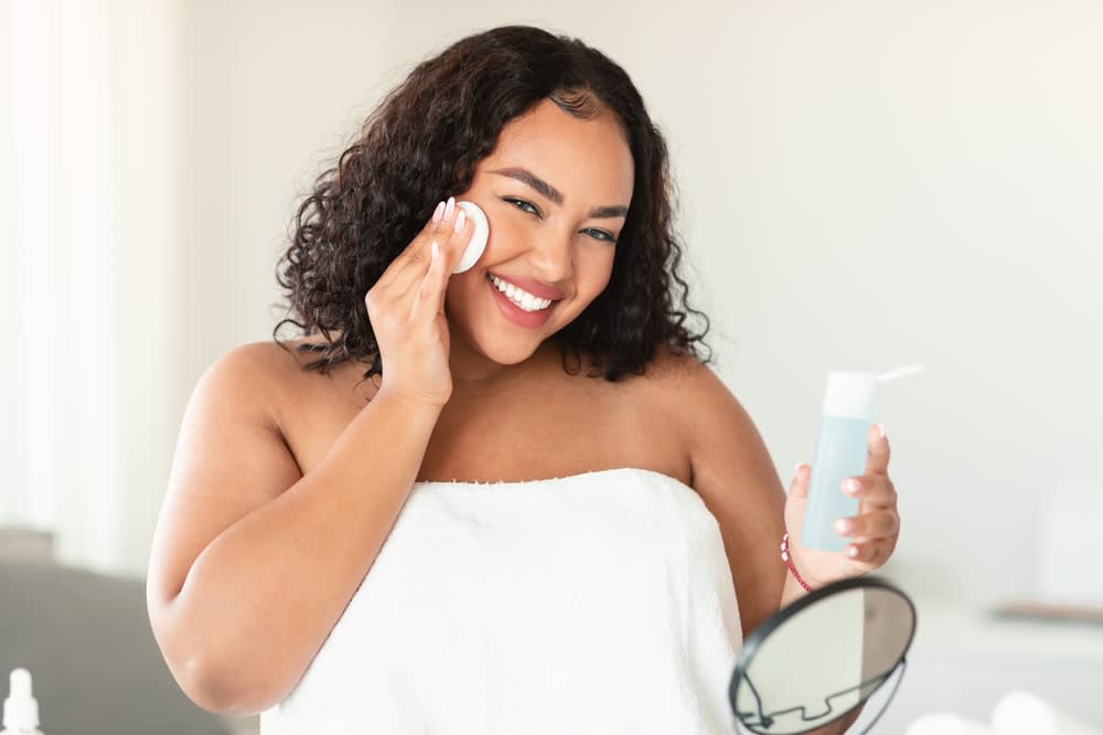 Imagem de mulher em frente ao espelho sorrindo enquanto aplica um produto de skincare na pele