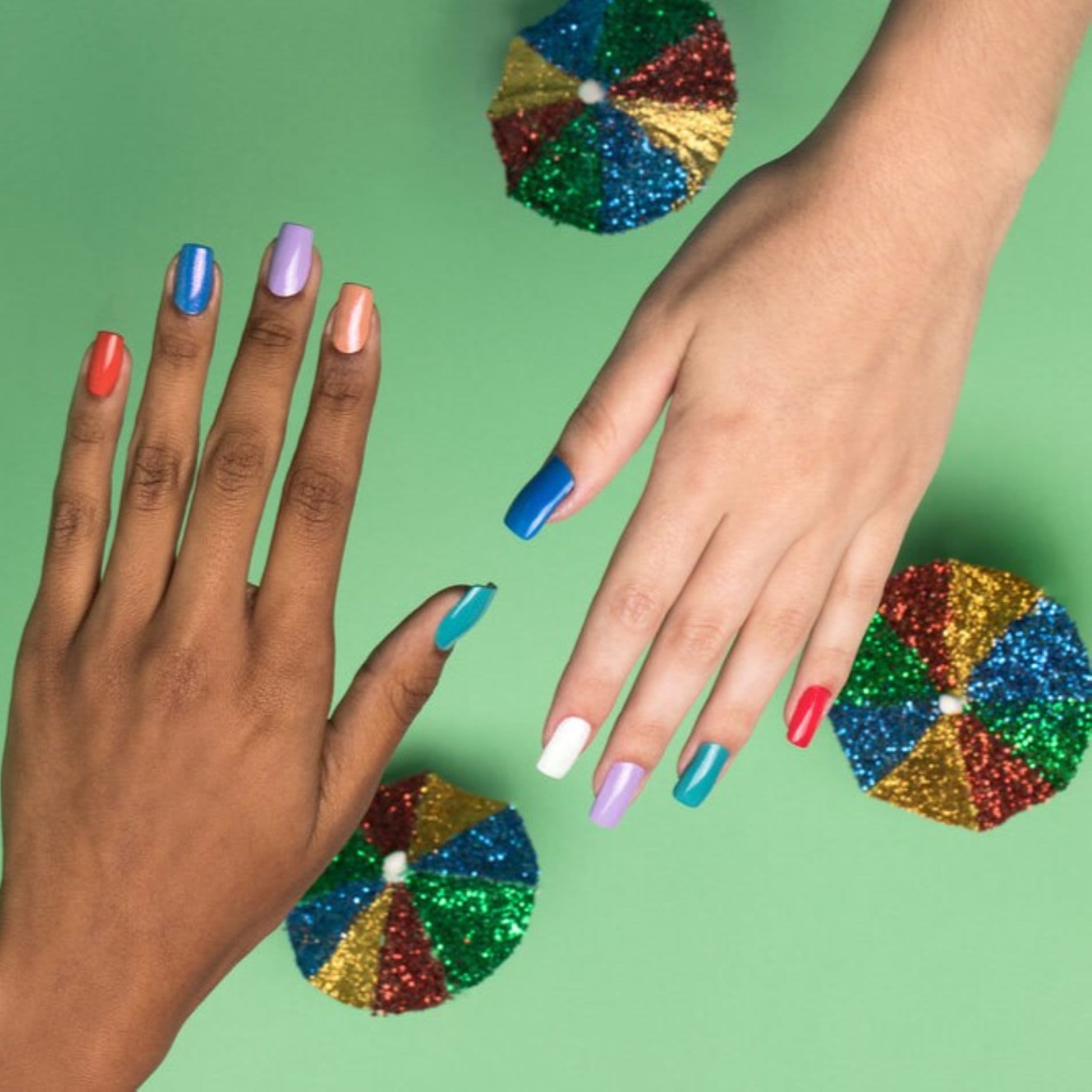 A imagem mostra duas mãos de pessoas diferentes com unhas coloridas, ao fundo está uma decoração de carnaval