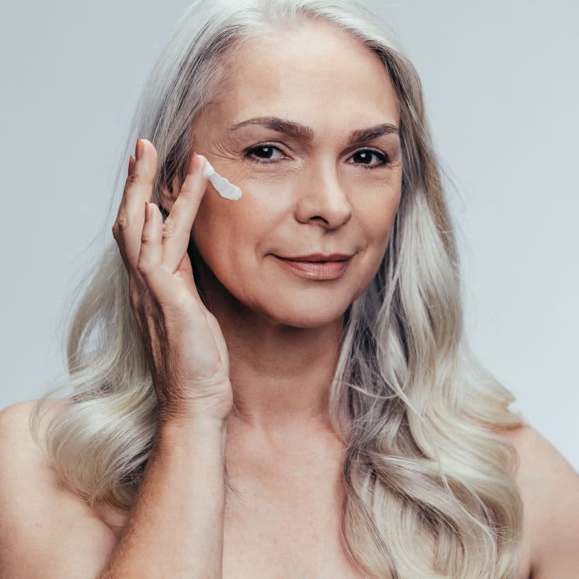 Mulher com cabelos grisalhos aplicando um creme hidratante no rosto, com expressão calma e confiante.
