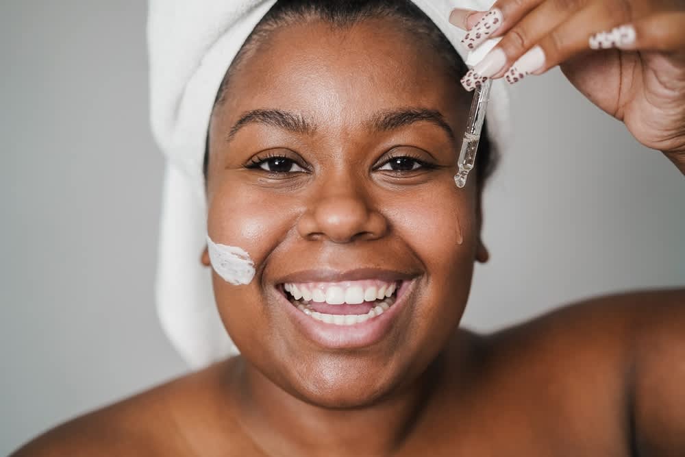 Mulher com toalha na cabeça fazendo rotina de skincare, ela está aplicando sérum facial e creme hidratante