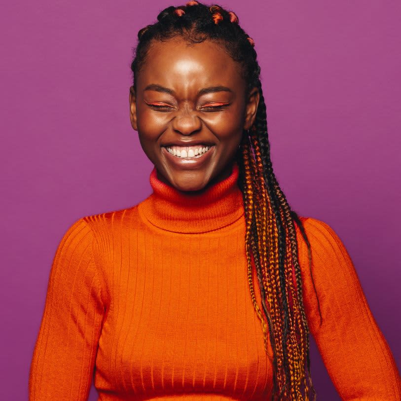 Mulher está sorrindo, ela tem a pele negra, usa tranças e usa roupas e maquiagem laranja