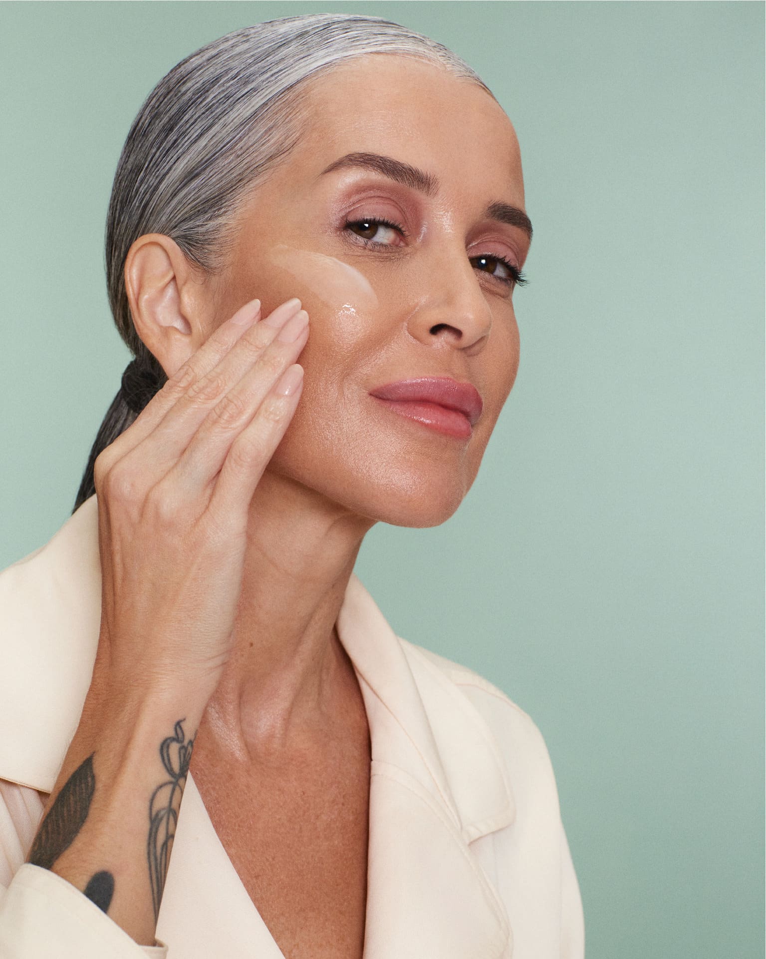 Mulher com cabelos grisalhos penteados para trás aplicando um produto cremoso no rosto com a ponta dos dedos, olhando para o lado. Uma tatuagem de folhas é visível em seu braço esquerdo.