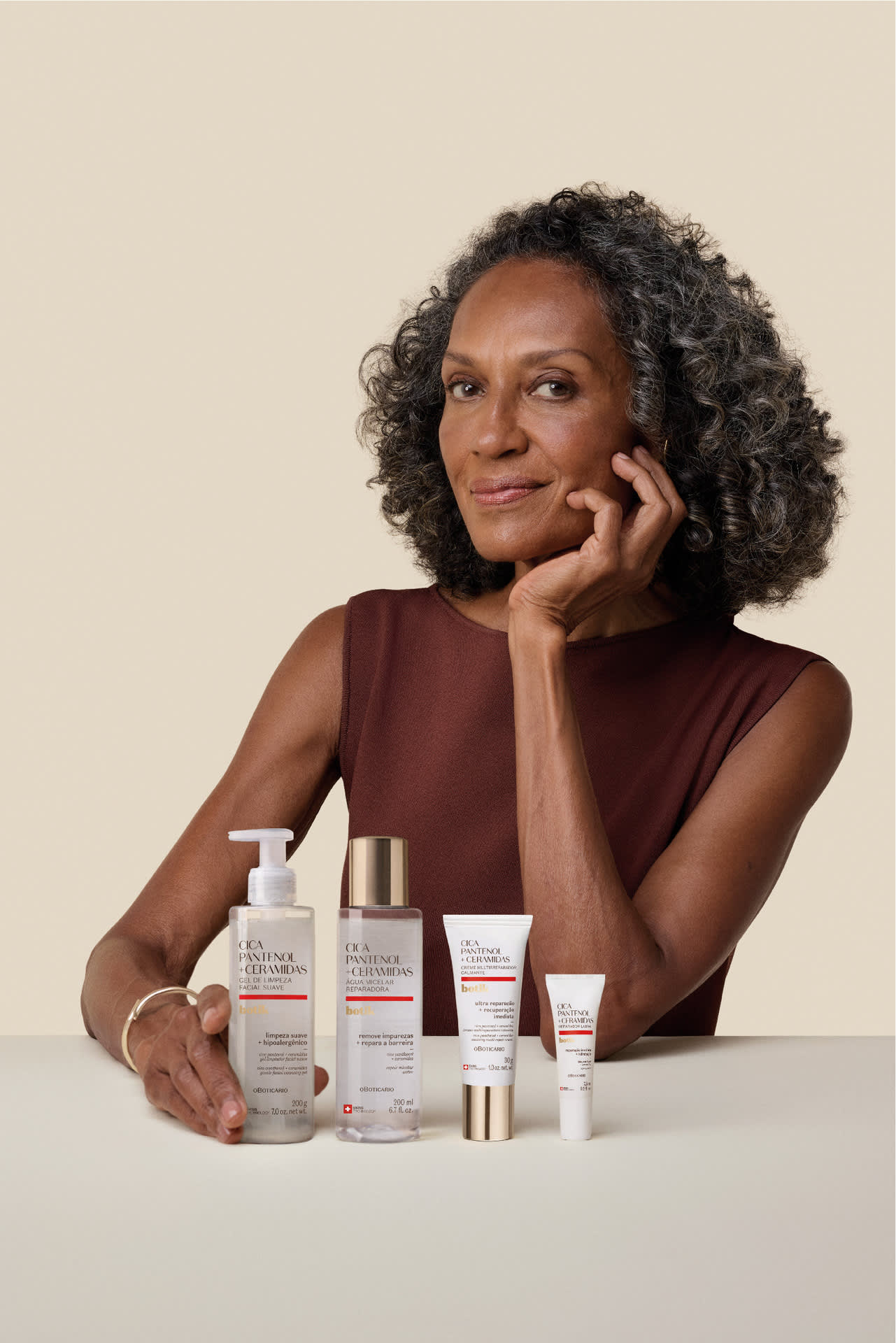 Mulher com cabelos grisalhos e cacheados sentada à mesa, com a mão no queixo e sorrindo, exibindo quatro produtos de skincare da linha Botik Pantenol + Ceramidas.