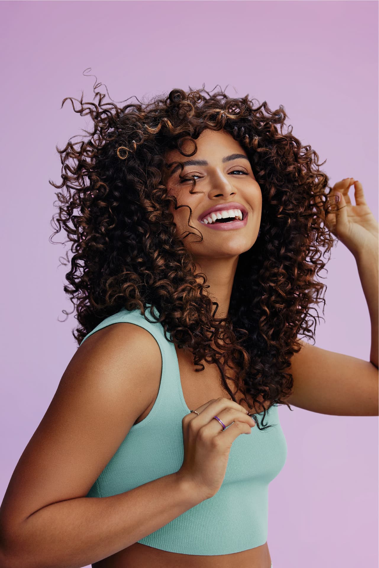 Uma mulher sorrindo, com cabelos cacheados volumosos e bem definidos na cor castanho claro, vestindo um top verde-água, em um fundo lilás.