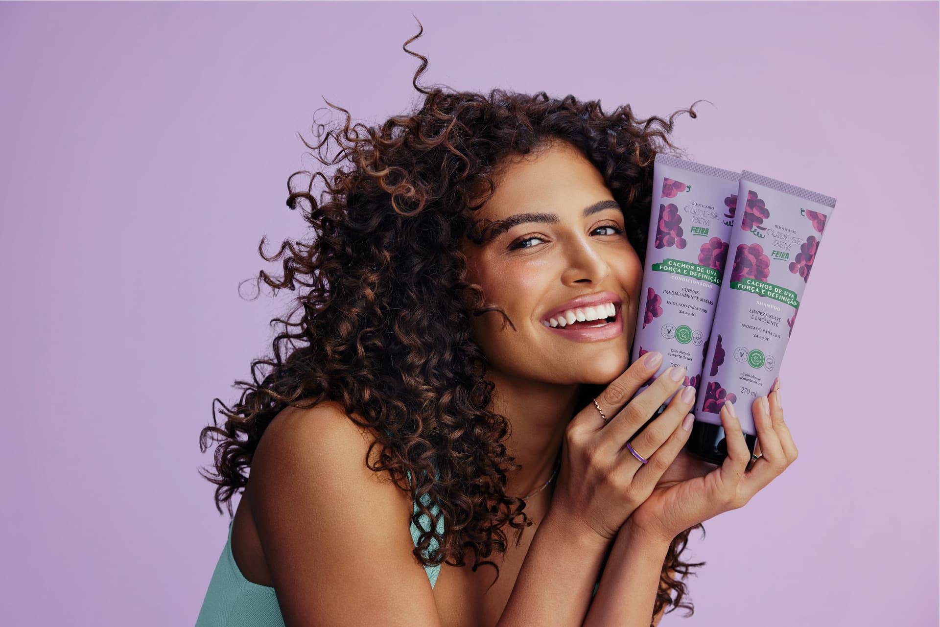 Uma mulher sorridente com cabelos cacheados volumosos, segurando dois produtos da linha Cuide-se Bem Cachos de Uva (shampoo e condicionador) em embalagens roxas com ilustrações de uvas, em um fundo lilás.