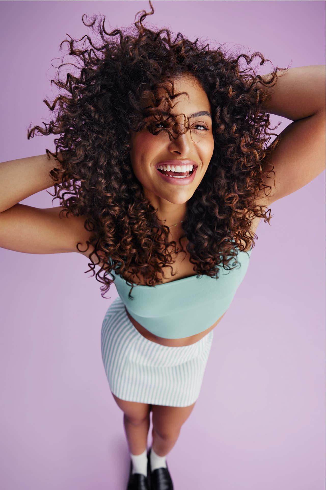 Uma mulher sorridente com cabelos cacheados volumosos e definidos, vestindo um top verde-água e uma saia listrada branca e cinza, em um fundo lilás. Ela está com as mãos na cabeça e olhando para cima