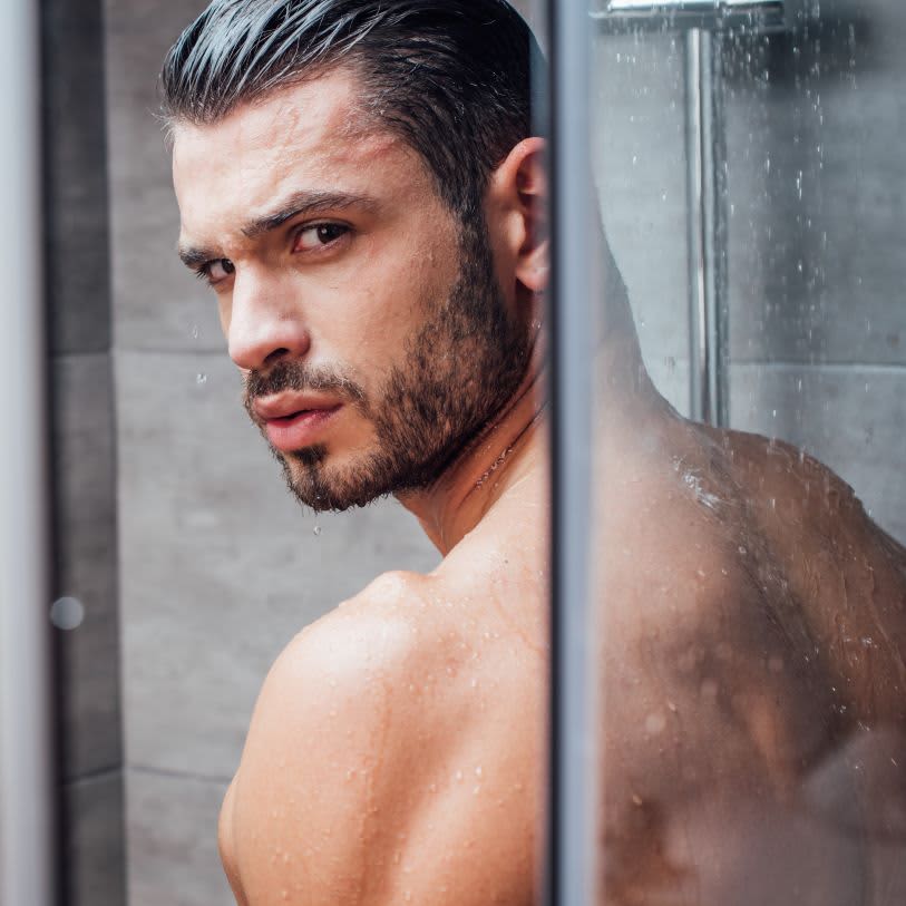 Homem moreno, molhado, com barba por fazer, olha para a câmera no chuveiro.