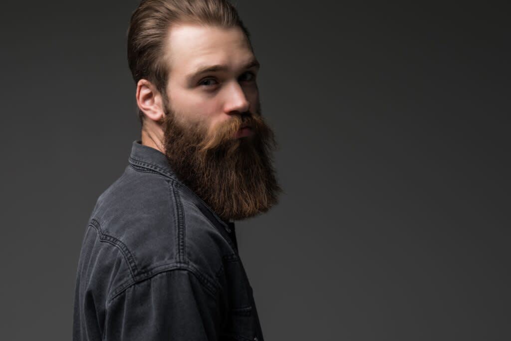 Homem em fundo cinza com barba longa e cabelo castanho. Veste uma camisa jeans também cinza.