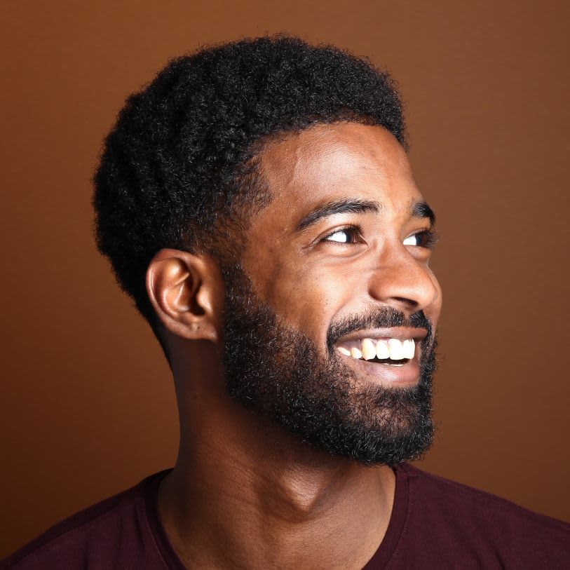 Homem sorridente com corte americano e barba. Ele olha para a direita, contra um fundo marrom.