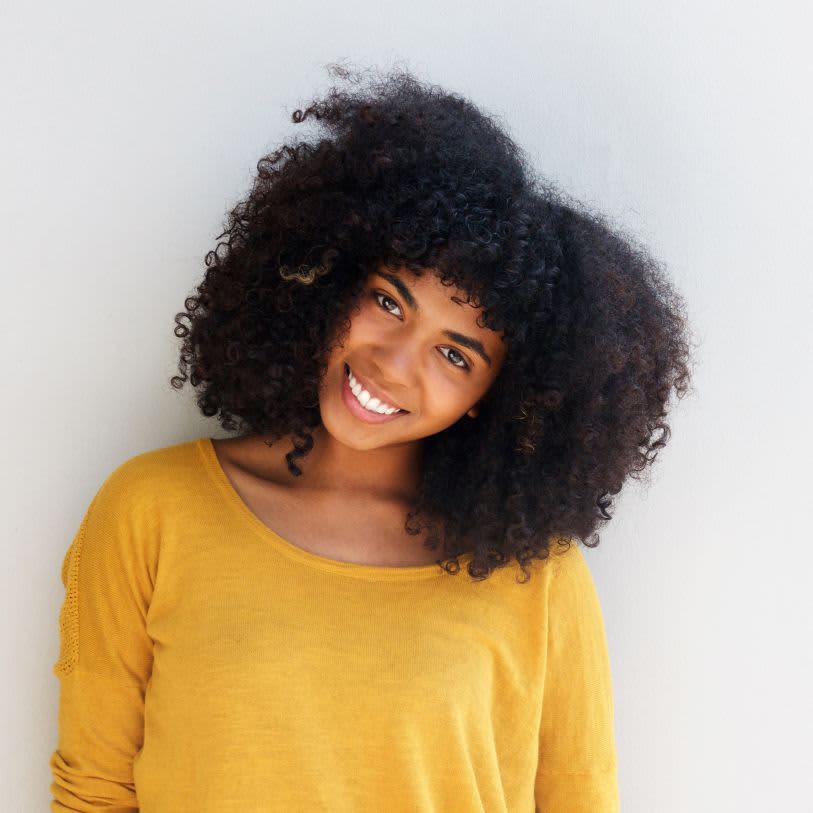 Mulher radiante com um deslumbrante cabelo crespo, vestindo blusa amarela e exibindo um sorriso autêntico em um ambiente minimalista.