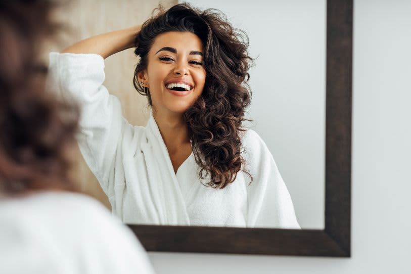 Mulher radiante de roupão branco sorrindo para o espelho, celebrando o volume e a beleza de seus cabelos cacheados.