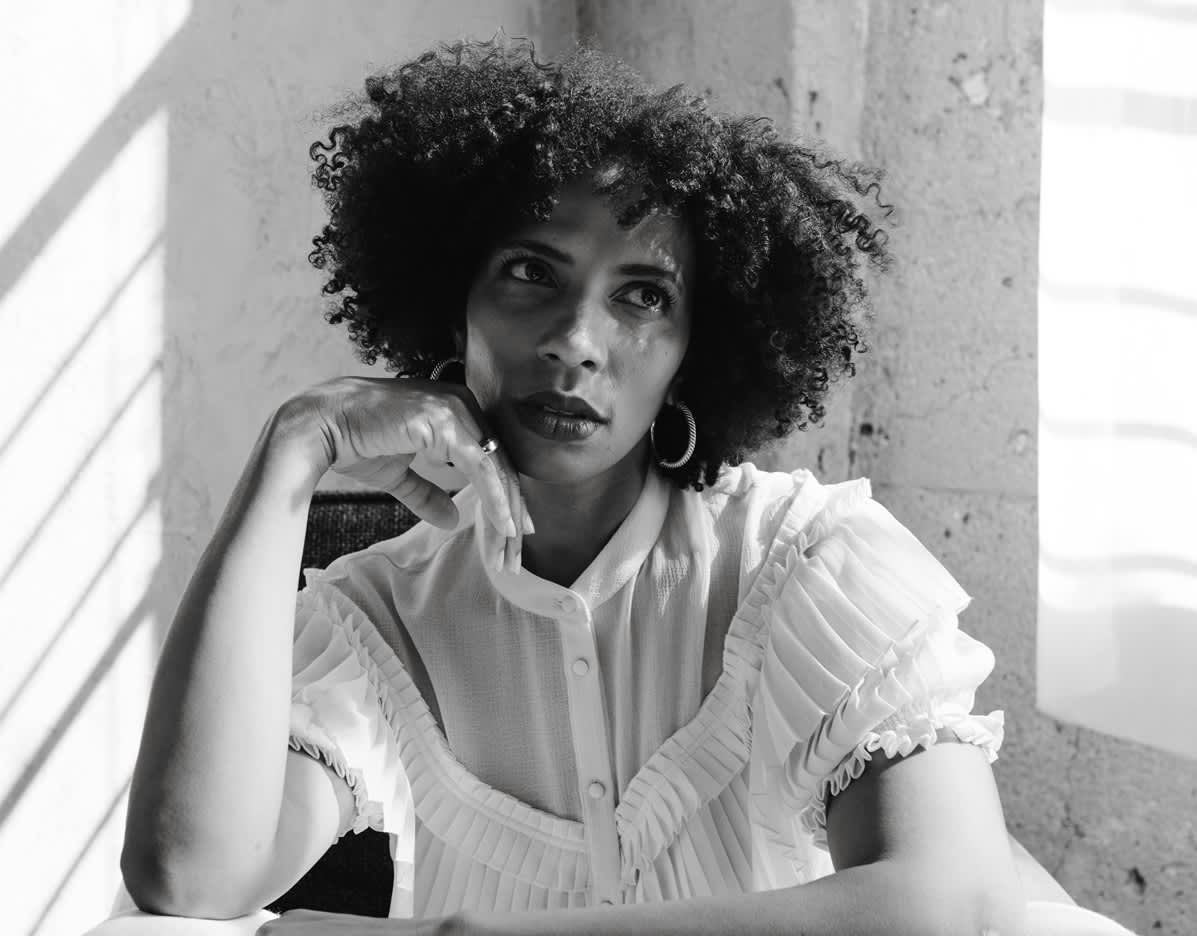 Mulher de cabelo crespo bem vestida com a mão apoiada no queixo. A foto está em preto e branco