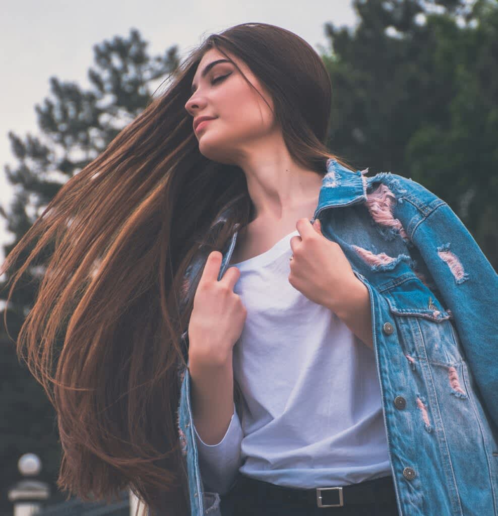 Mulher de cabelo longo castanho jogando ele de lado. Ela está de jaqueta jeans azul e camiseta branca.