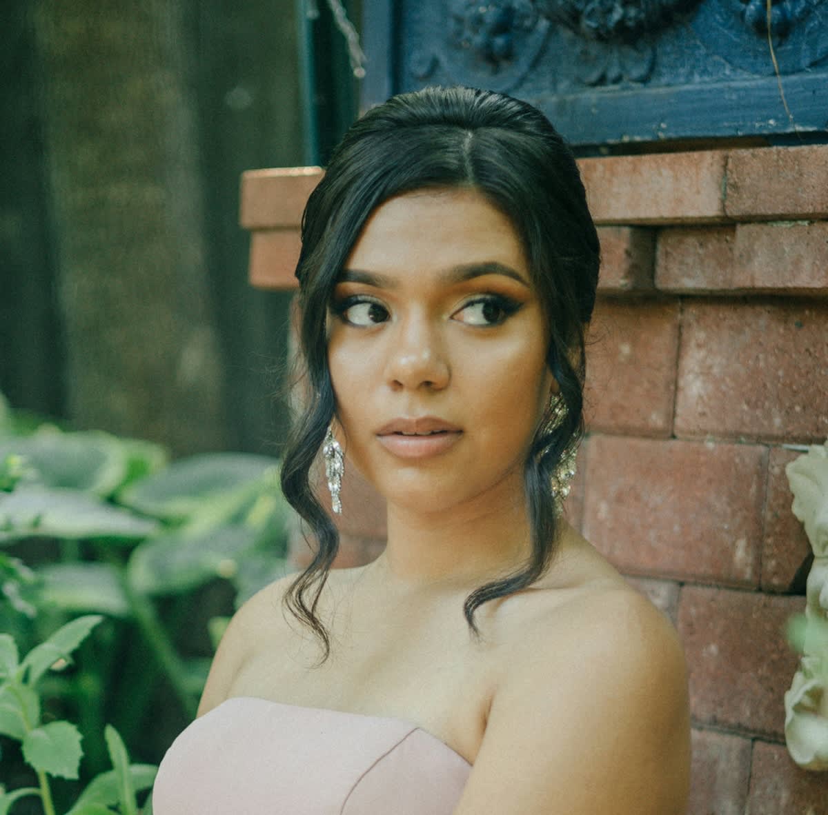 Mulher jovem de cabelos presos, com brincos grandes e maquiagem de festa. Usa um vestido tomara que caia rosa bebê.