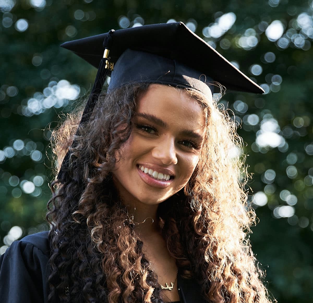 Mulher negra clara, jovem com cabelos longos cacheados. Ela está sorrindo para a foto e veste beca e chapéu de formatura, ambos de cor preto.