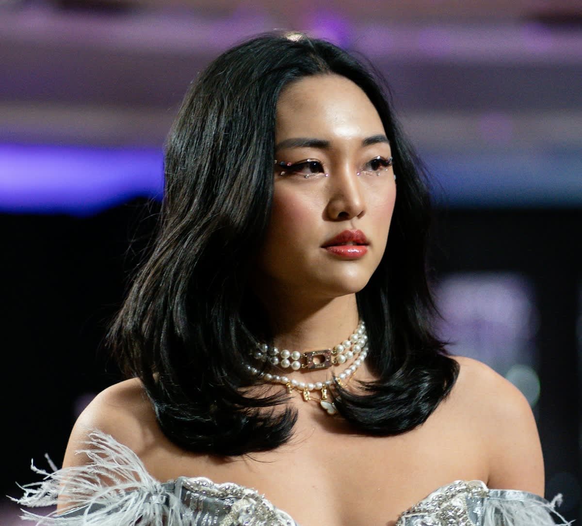 Mulher com traços asiáticos com maquiagem de formatura com vários strass. Ela tem cabelo médio preto e liso. Usa um colar de pérolas.