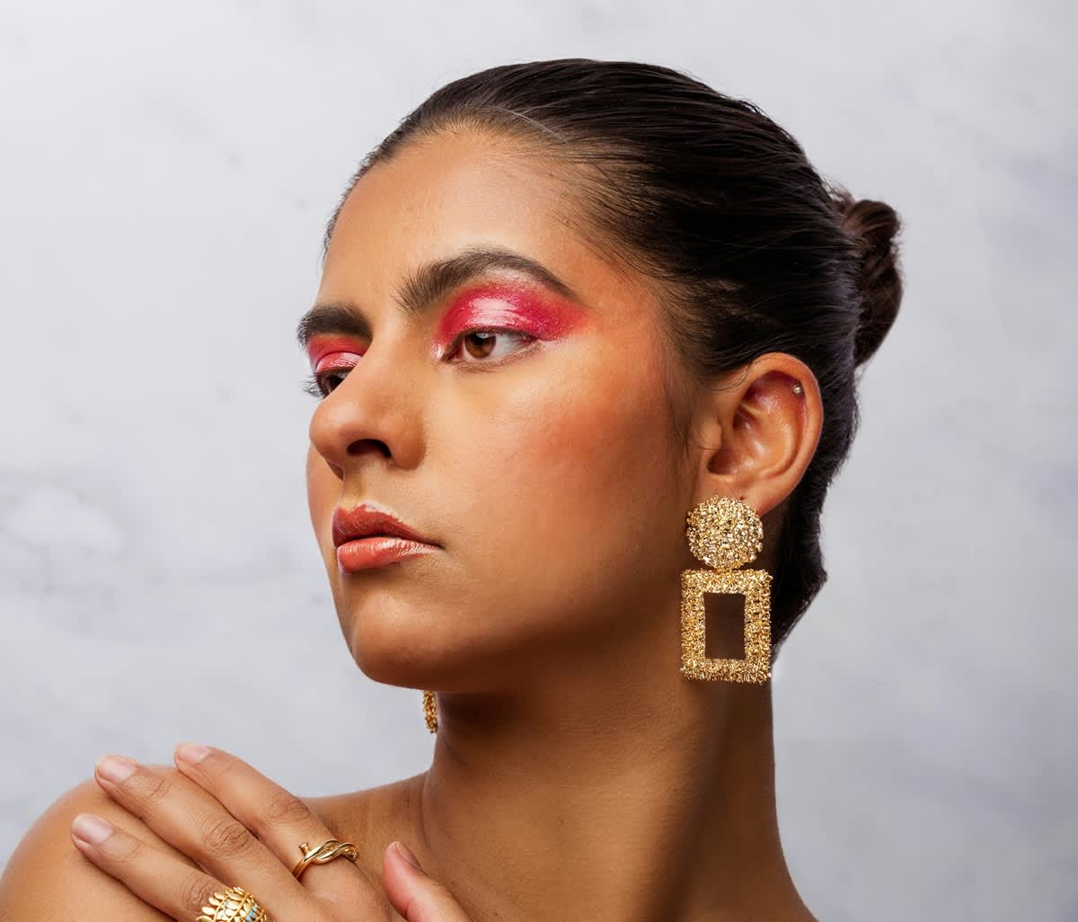 Mulher branca com coque e brincos dourados. A maquiagem que ela usa nos olhos tem um tom de pink bem vivo e os lábios tem gloss.