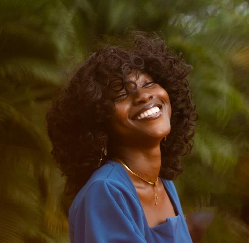 Mulher de pele retinta sorrindo de olhos fechados. Seu cabelo cacheado está bem definido e ela tem uma franja. Usa um vestido azul de mangas compridas e está em uma local externo com bastante verde em volta.