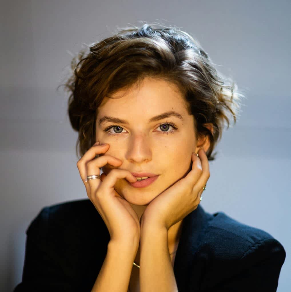 Mulher de cabelos curtos e mãos no rosto olha para a câmera com leve sorriso.