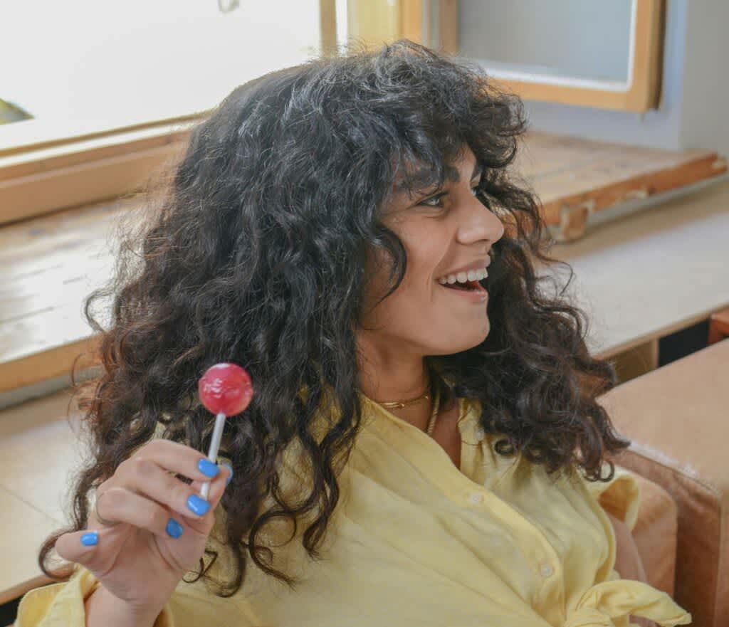 Mulher de cabelos ondulados longos com franja. Ela está de camisa amarela e sorri sem olhar para câmera. Na mão, segura um pirulito.