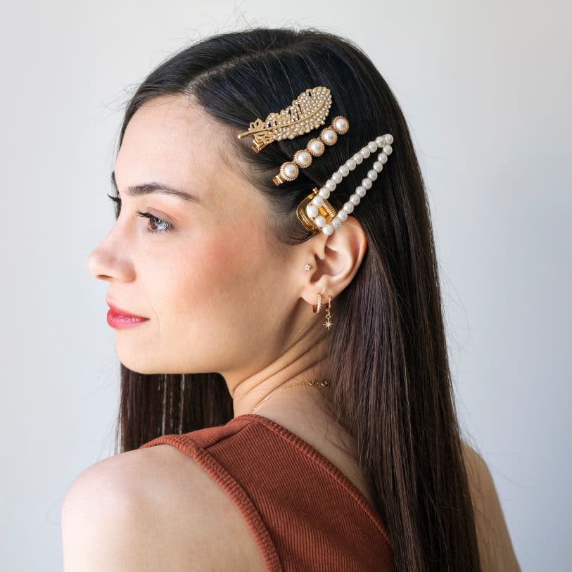 Jovem de perfil com cabelos lisos e escuros, adornados por três presilhas luxuosas de pérolas e brilhos, trazendo um toque clássico e moderno.