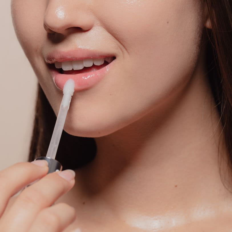 Close do rosto de uma mulher aplicando gloss labial transparente. Foco nos lábios, dentes brancos e pele com acabamento natural e viçoso.