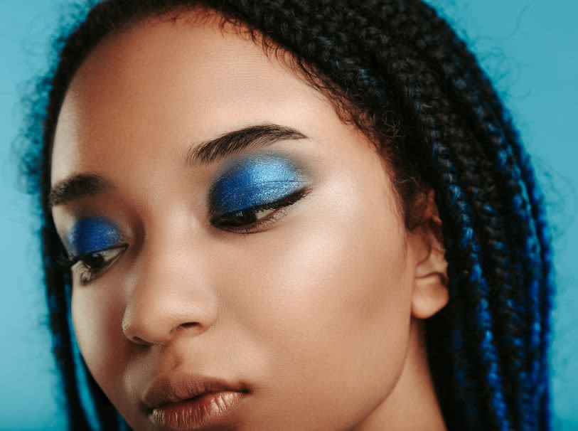 Close de uma mulher negra com sombras azuis vibrantes e tranças escuras com fios azuis. Pele iluminada e fundo azul-claro.