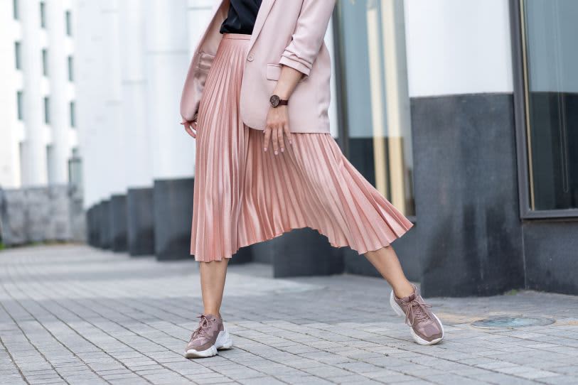 Mulher usando blazer e saia plissada rosa metálica com tênis esportivos. Look moderno em ambiente urbano com fundo de colunas brancas.