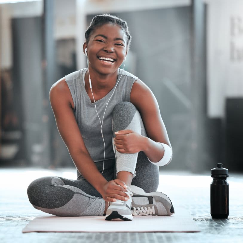 Apresenta uma mulher negra sentada em um colchonete no chão da academia, sorrindo alegremente para a câmera. Ela usa fones de ouvido brancos conectados ao celular, roupas esportivas cinzas e há uma garrafa de água preta ao seu lado.