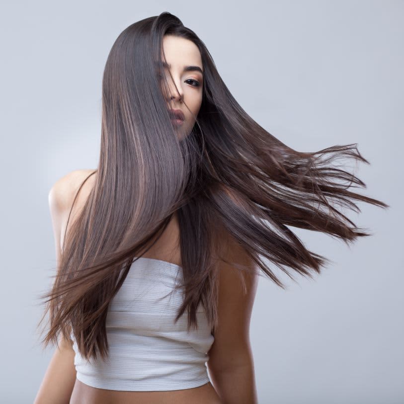 Mulher com cabelos longos, lisos e castanhos em movimento ao vento, usando um top branco frente a um fundo cinza claro.