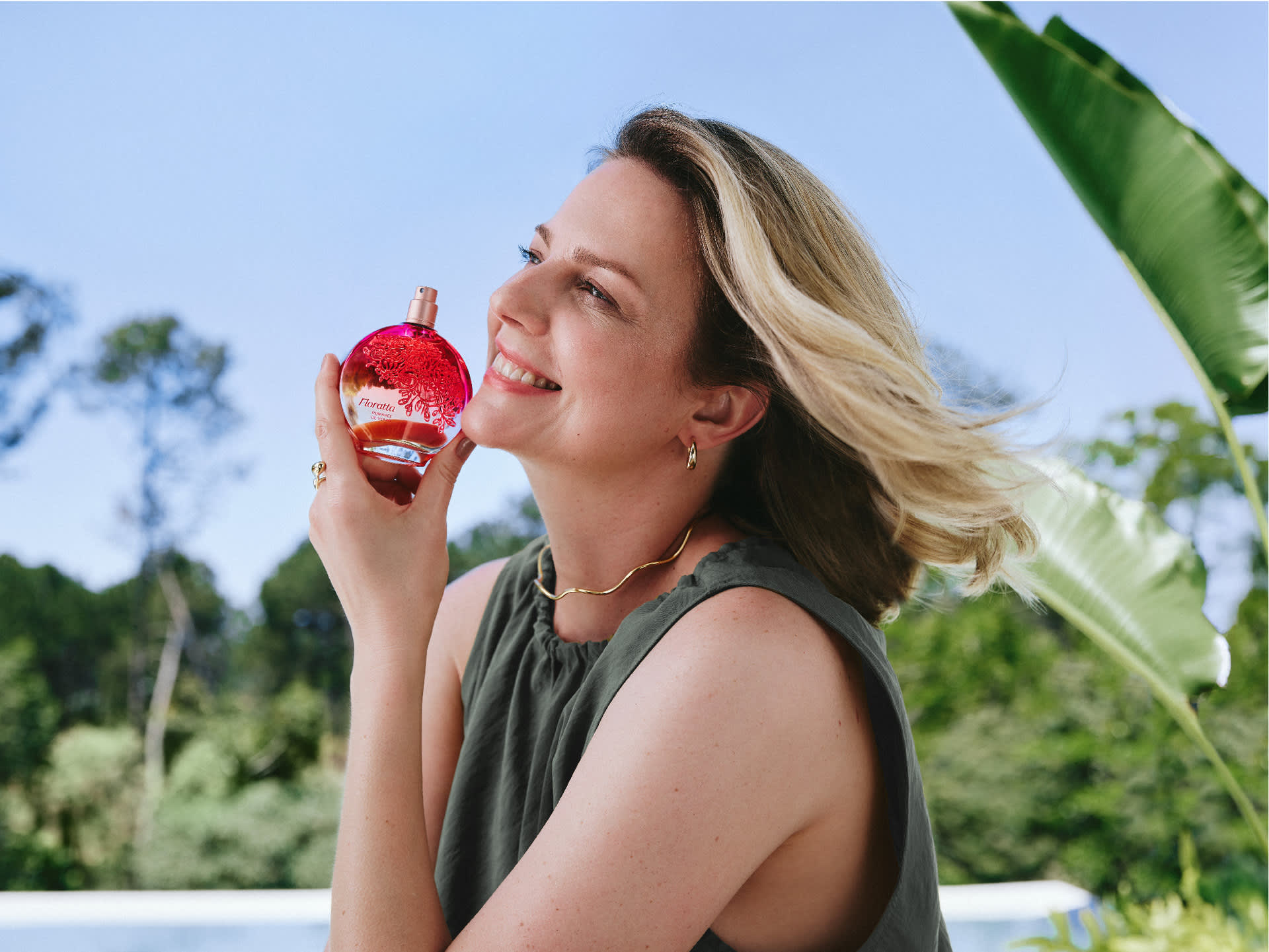 sorridente ao ar livre segura um frasco de Floratta Romance de Verão, com folhagem tropical verde e céu azul ao fundo.
